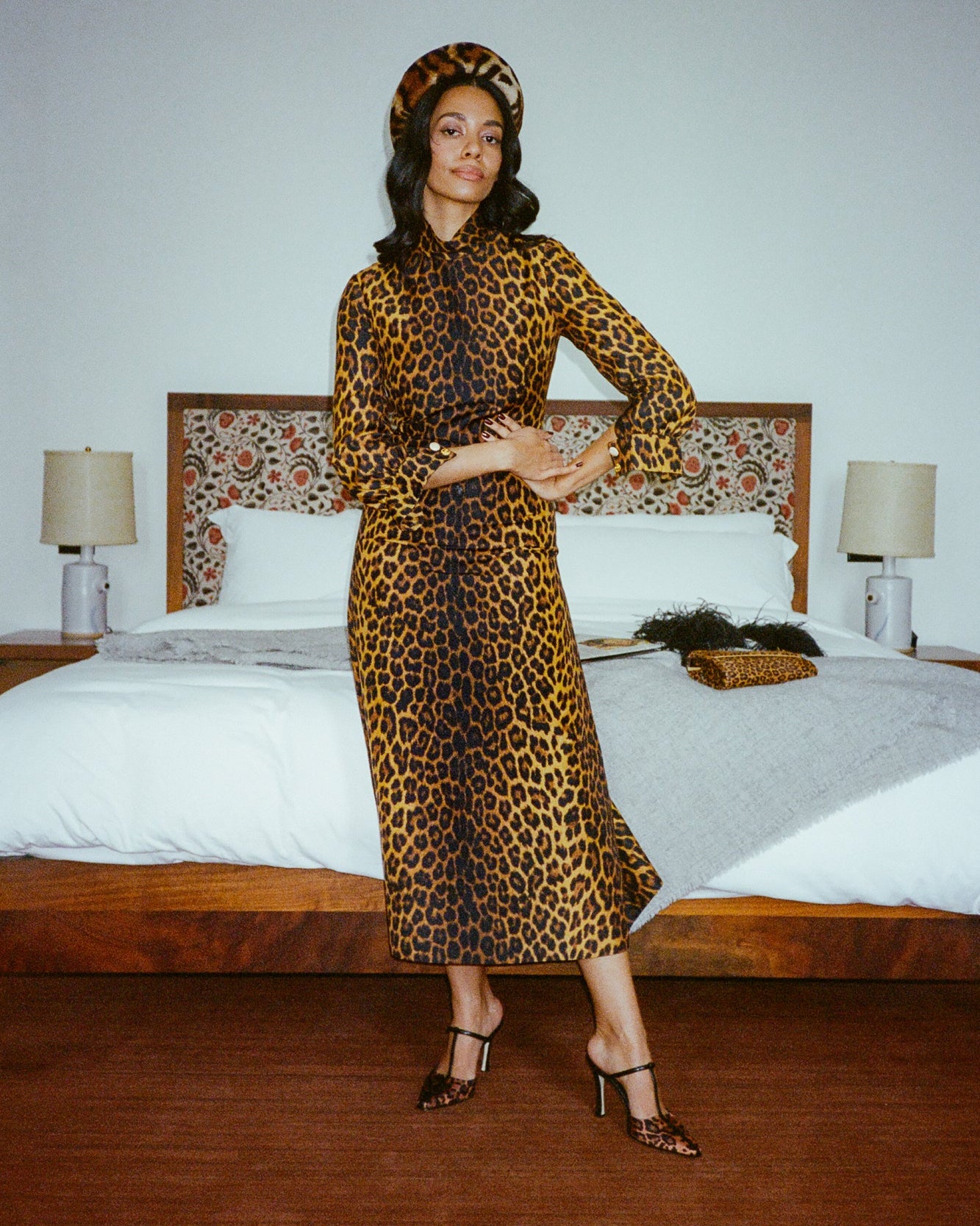 A woman stands in a bedroom wearing a leopard-print midi dress with a matching beret and black strappy heels. The dress has a brown and black animal print on a tan background, and the room features a wooden bed frame, white bedding, and bedside lamps.