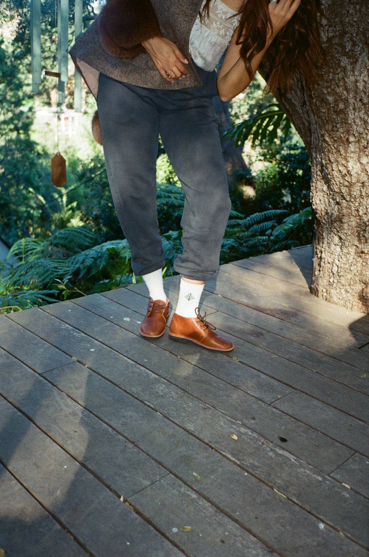 A person standing on a wooden boardwalk outdoors under a tree, wearing gray trousers, white socks, and brown leather shoes. The photo is taken from a low angle and appears slightly tilted.