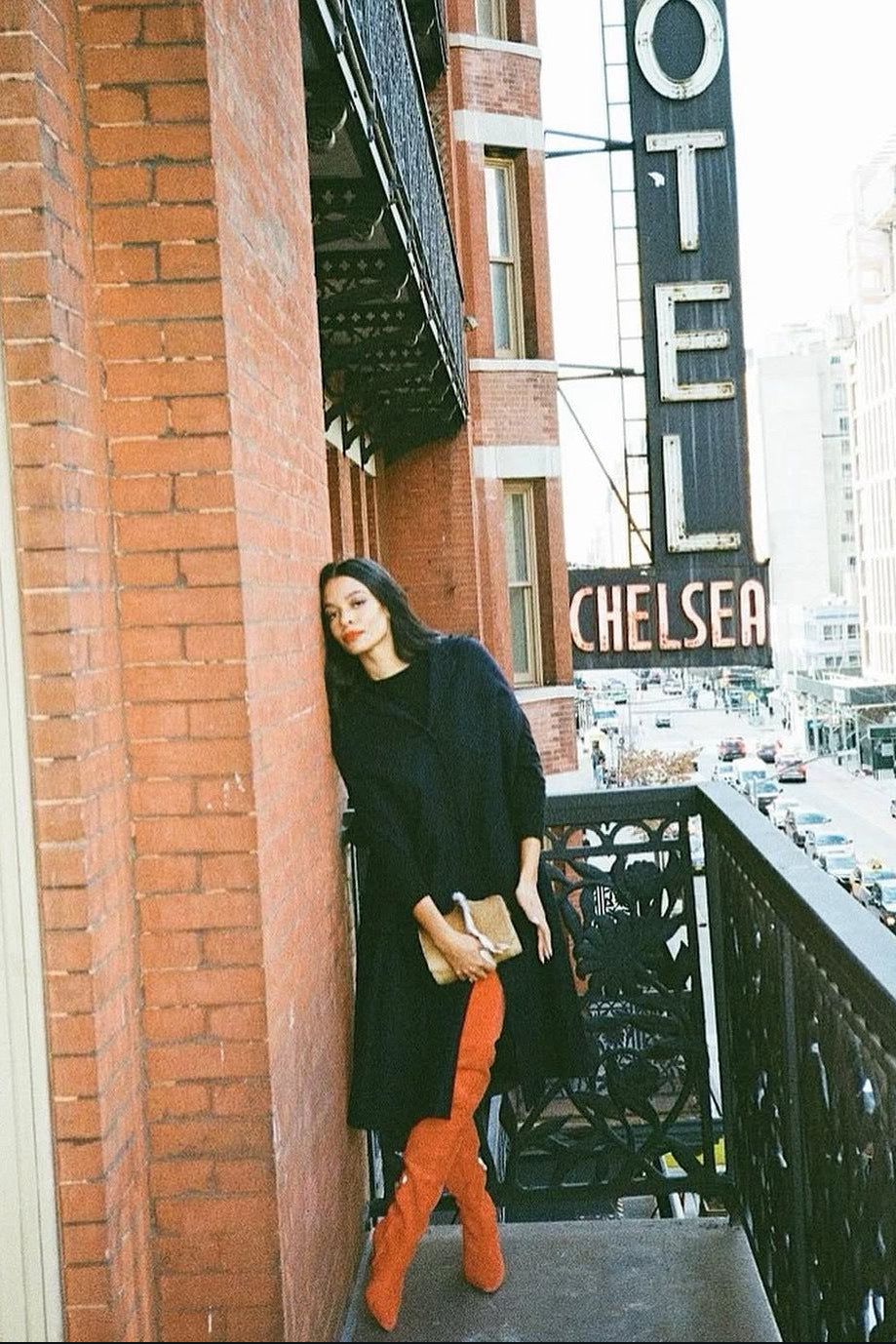 A woman stands on a red brick building balcony in an urban city setting, wearing a long black coat and bright rust-orange over-the-knee boots, holding a small beige clutch.