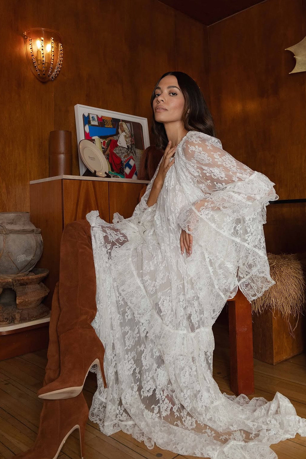 A woman wearing a white lace maxi dress and brown suede over-the-knee boots poses in a rustic, warm-toned room with wood-paneled walls and decorative objects.