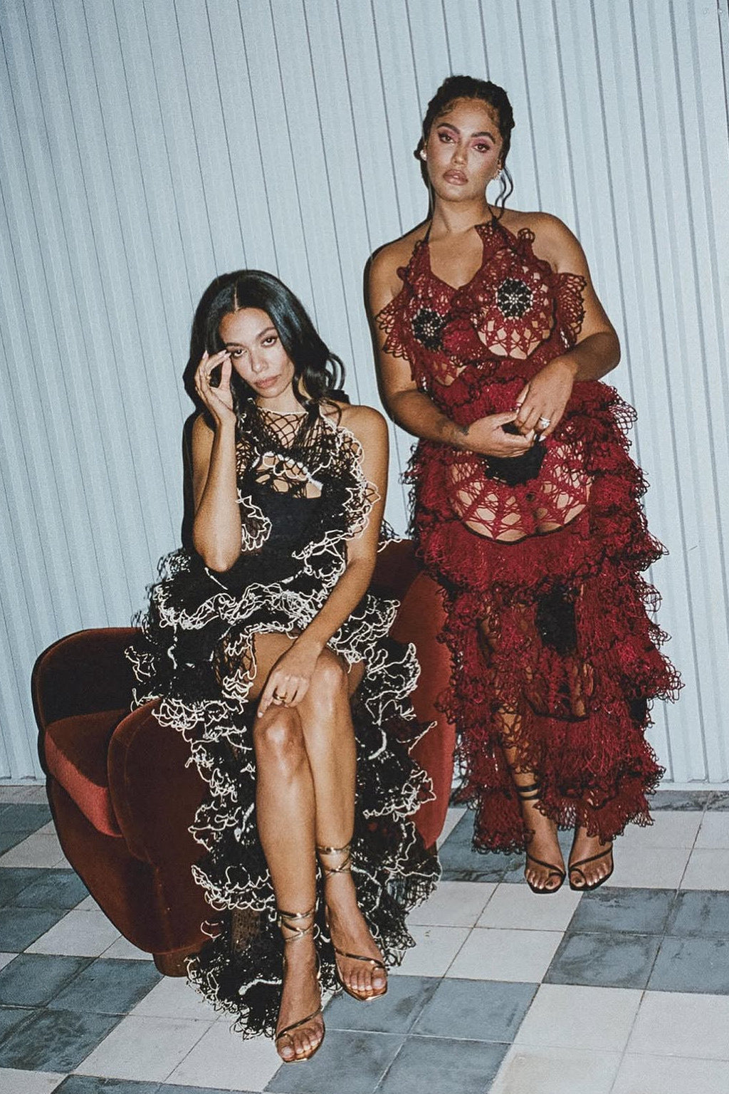 two women wearing long lace gowns and strappy heeled sandals. One is seated and the other is standing.