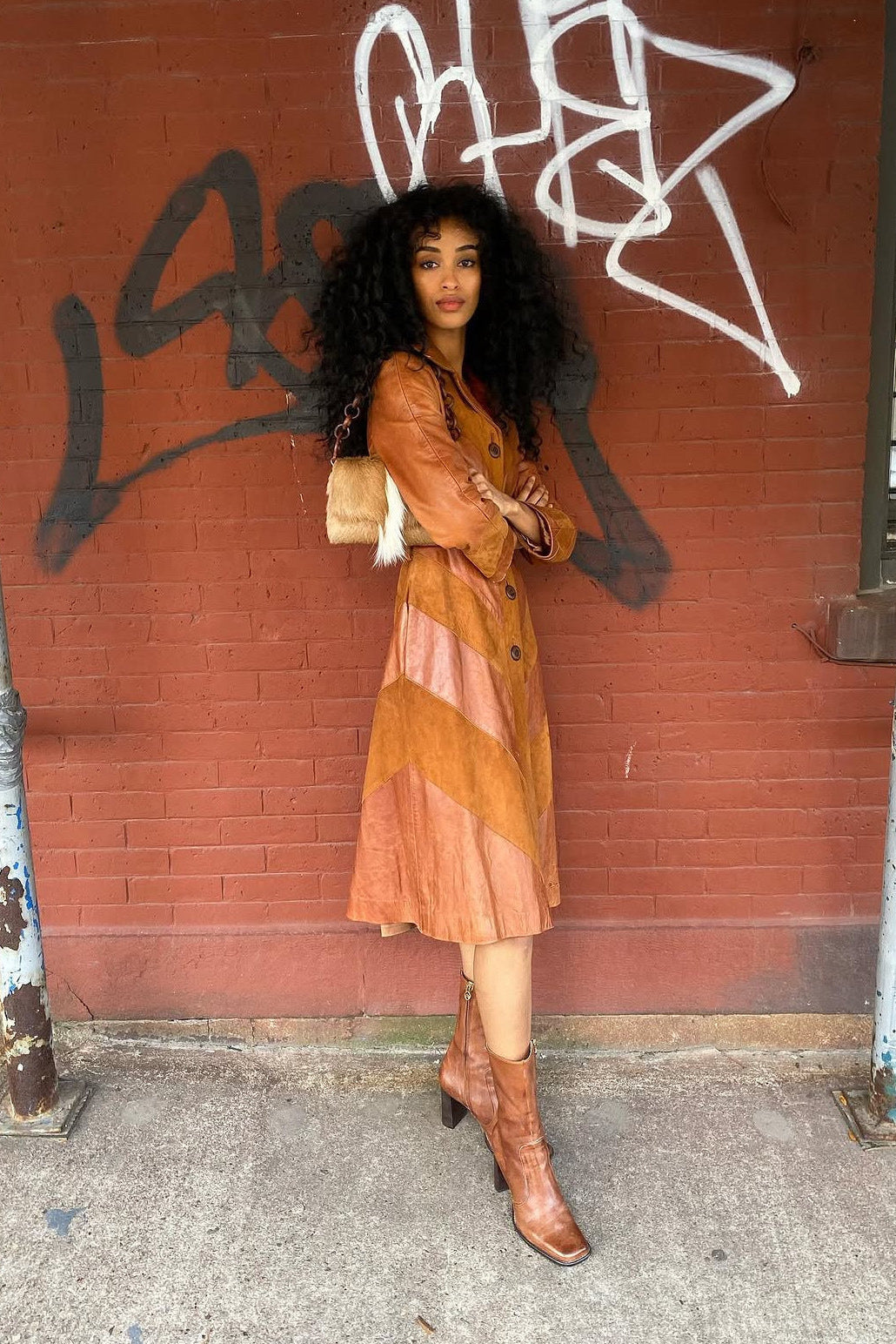 A woman stands on a city sidewalk in front of a red brick wall with graffiti, wearing a rust-orange leather trench coat with diagonal panels and matching brown leather knee-high boots.