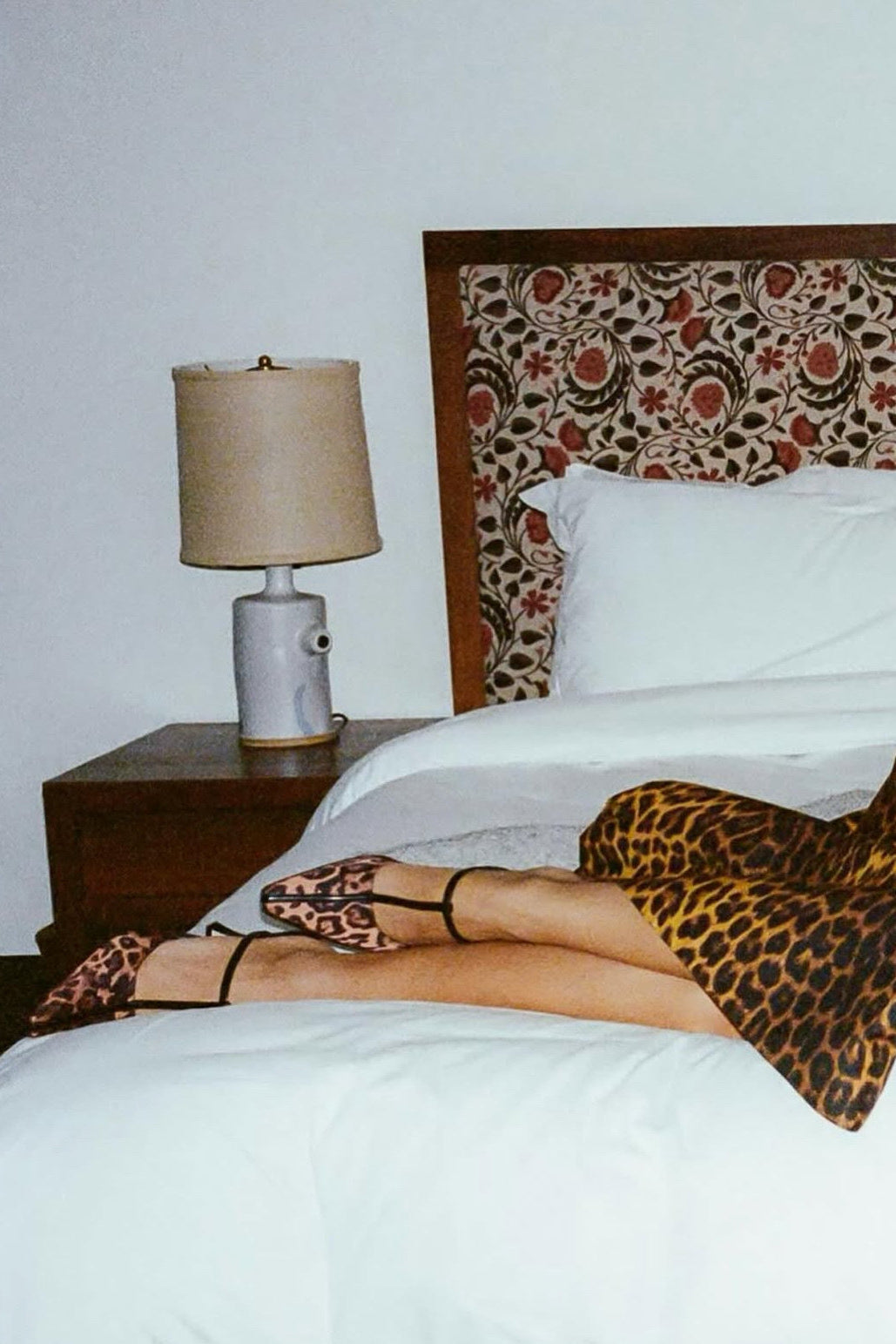 A person reclining on a bed wearing a leopard-print dress and matching leopard-print strappy heels, with a patterned headboard, white bedding, and a bedside lamp visible.