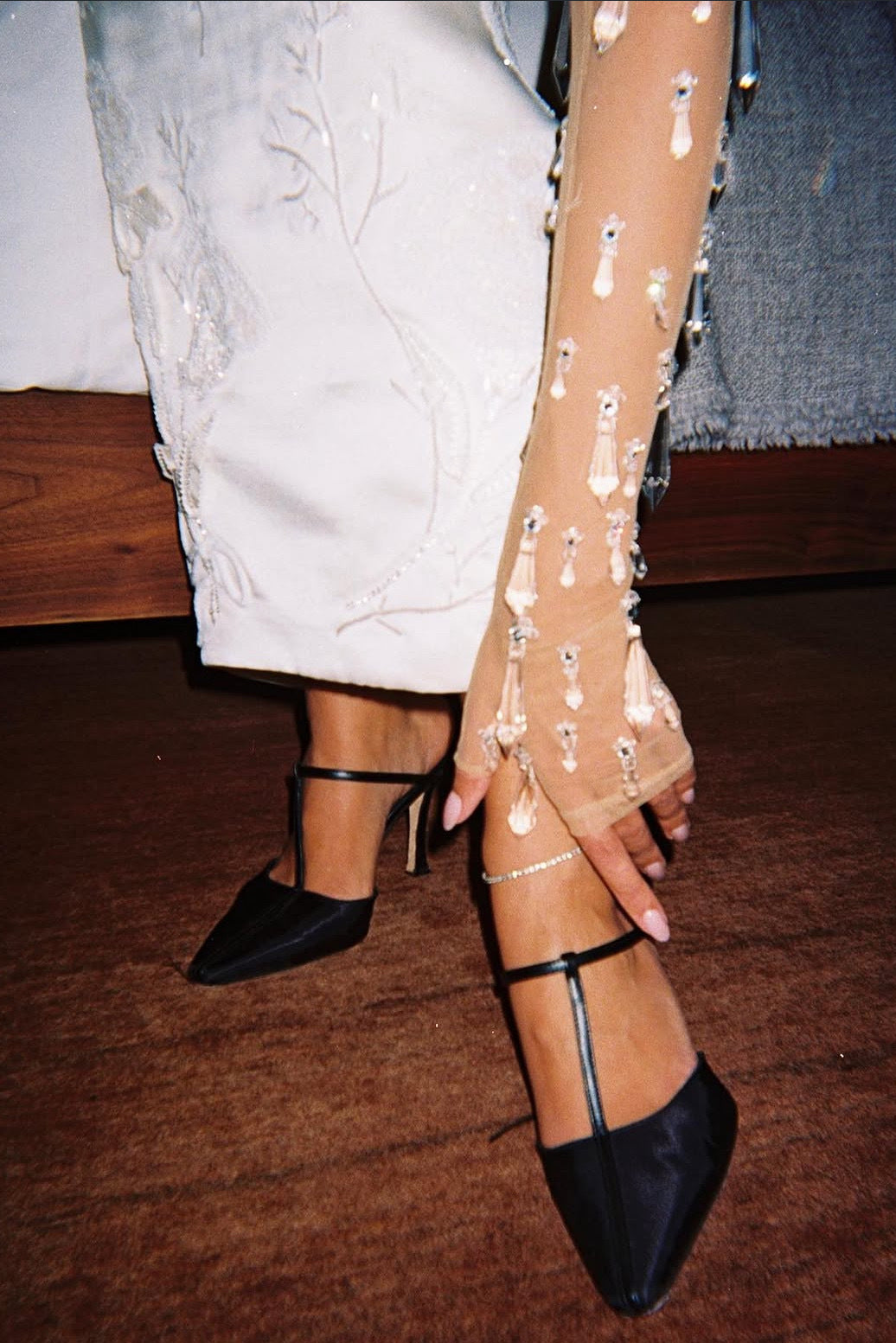 A close-up lifestyle shot of a person wearing black satin pointed-toe high heels with ankle straps, paired with a white embroidered dress and sheer beige arm warmers decorated with white beadwork.