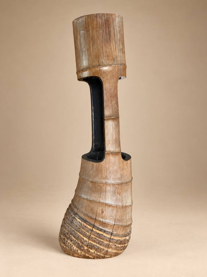 A tall, sculptural wooden vase or decorative vessel with a flared textured base, a narrow neck, and a cylindrical top section, shown against a plain beige studio background.
