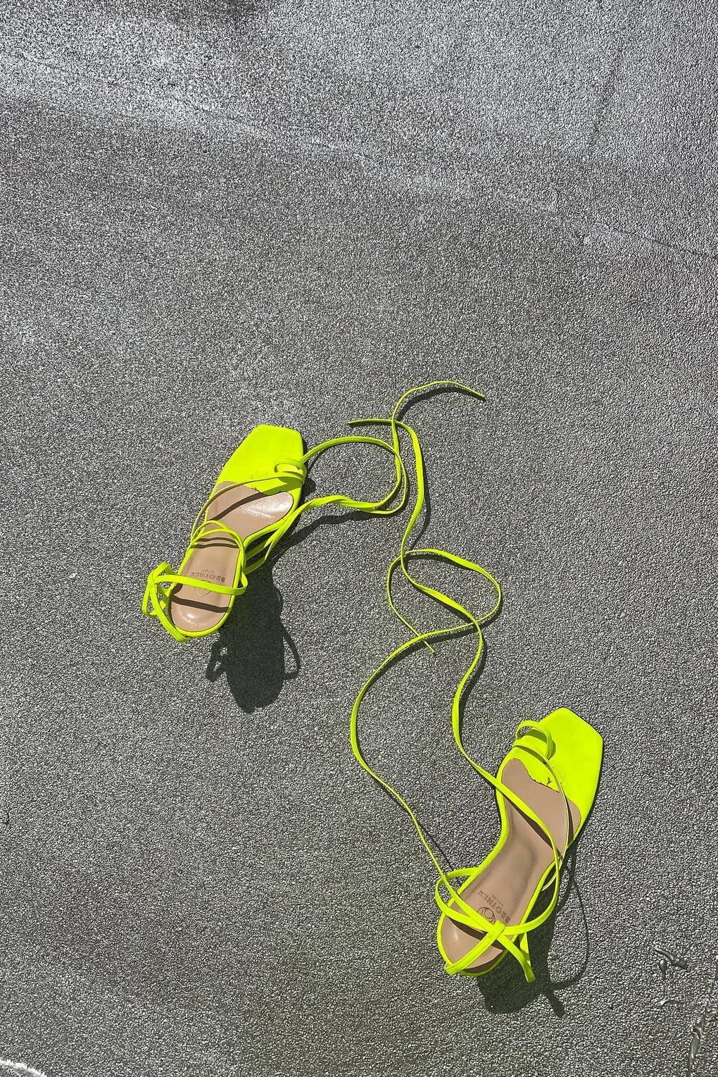 A pair of neon yellow strappy high-heeled sandals with square toes, laid out on a gray concrete surface.