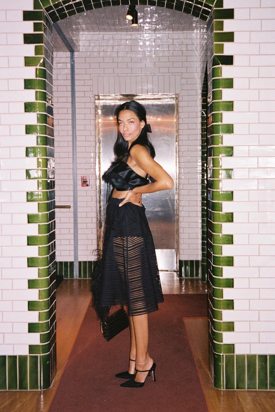 A woman posing in a black strapless satin top and a sheer black pleated midi skirt, holding a black handbag, set against a tiled hallway with green and white tiles.