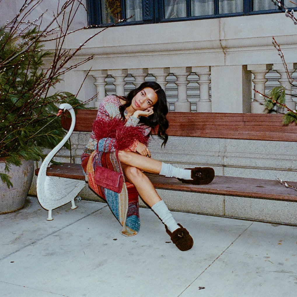 A woman sits on a wooden bench in an outdoor courtyard setting, wearing a colorful patterned coat, white socks, and dark loafers. The scene is framed by large potted plants and a stone wall in the background.