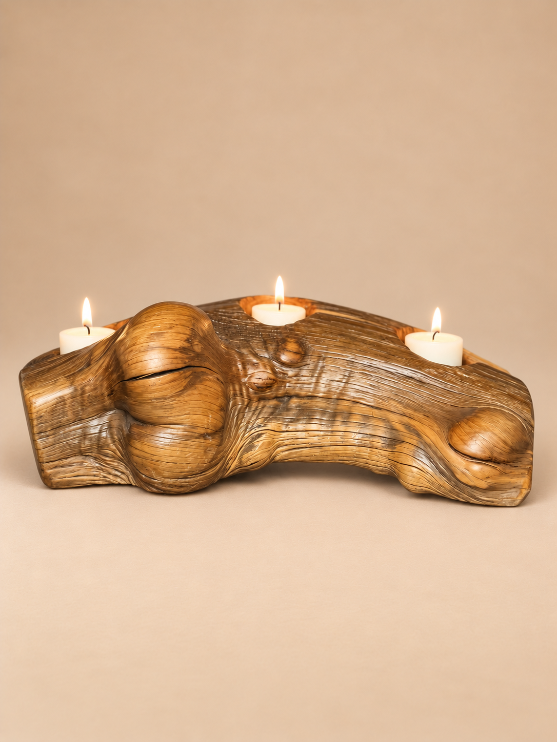 A mid-century modern carved teak wooden candle holder shaped like a tree trunk, holding three lit white pillar candles, set against a solid beige background. Burled wood with natural striations