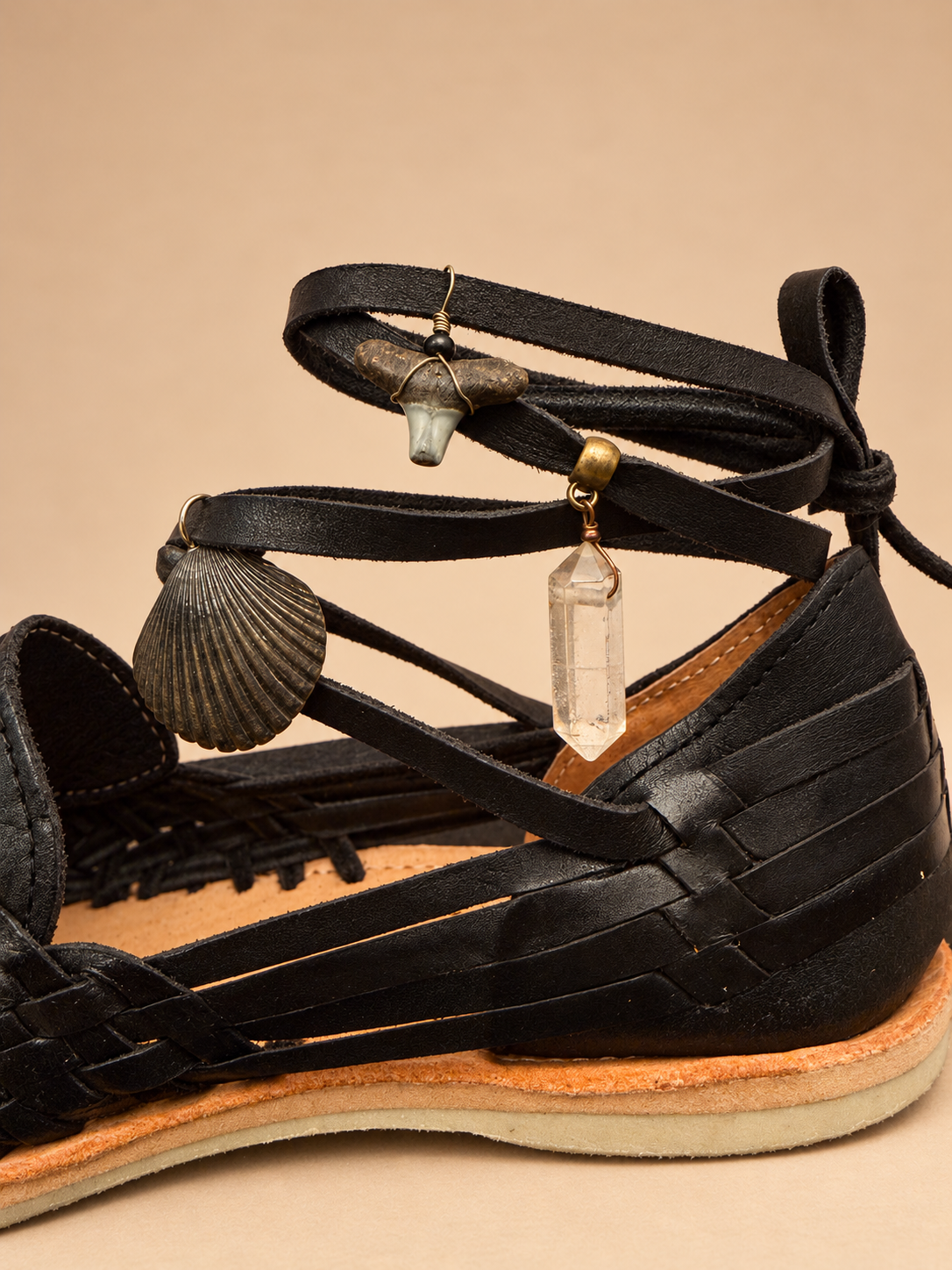 Candy Huaraches 013 in black woven leather, close-up of the scallop shell, shark tooth and clear quartz charms on the ankle laces.