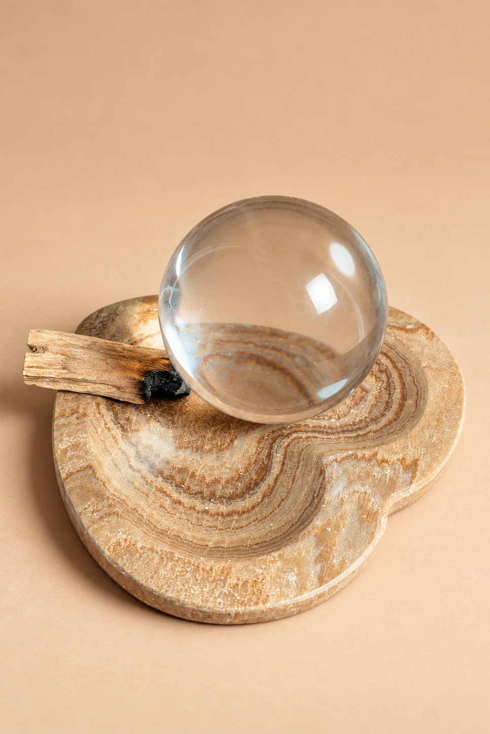 A clear glass crystal ball resting on a round, organic-shaped beige stone tray with swirling natural patterns, accompanied by a small piece of driftwood, set against a solid warm beige background.