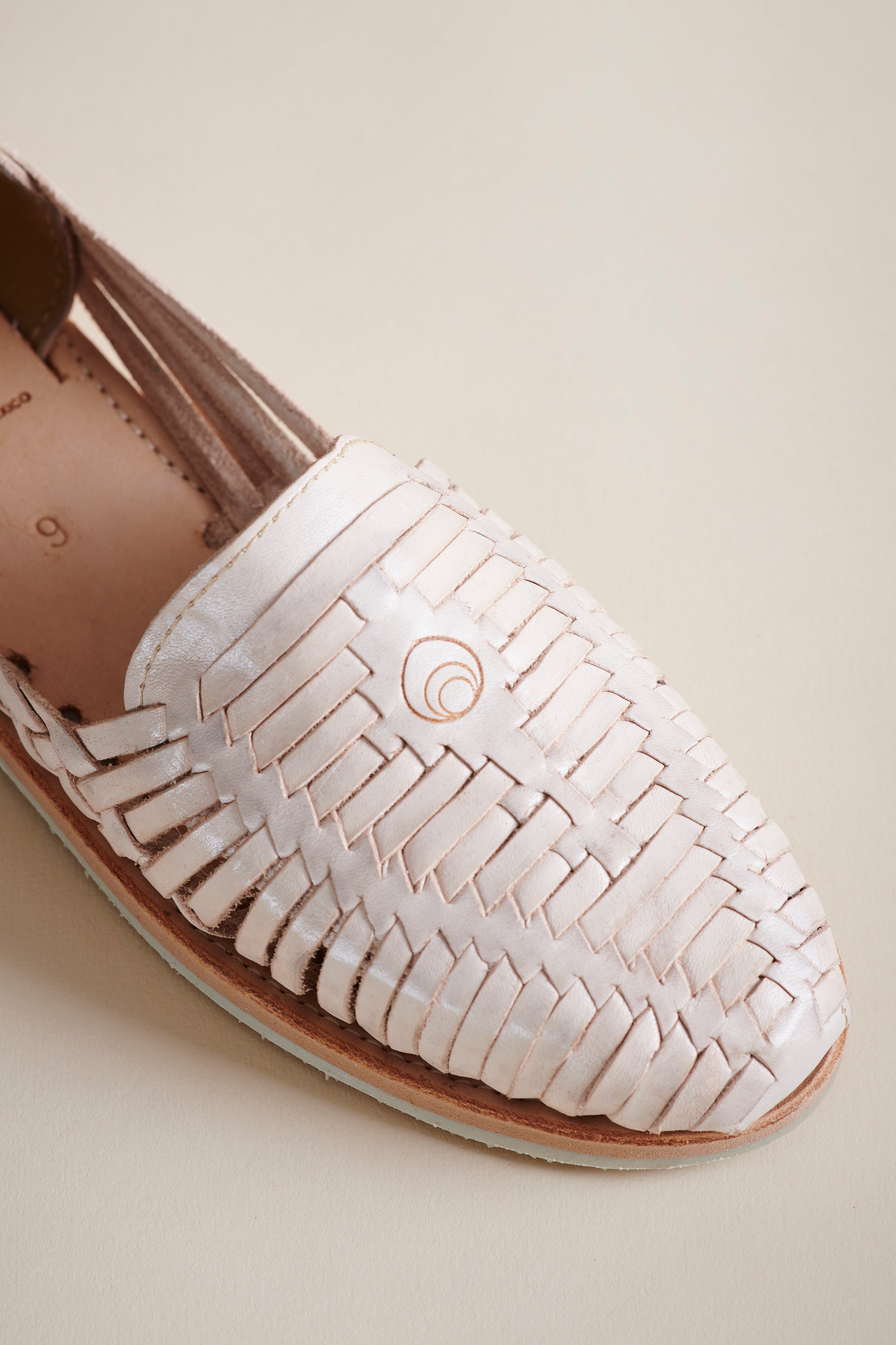 A close-up of a woven leather braided flat in a pale pink color, showing the detailed strap pattern and a small circular logo on the side, set against a solid beige background.