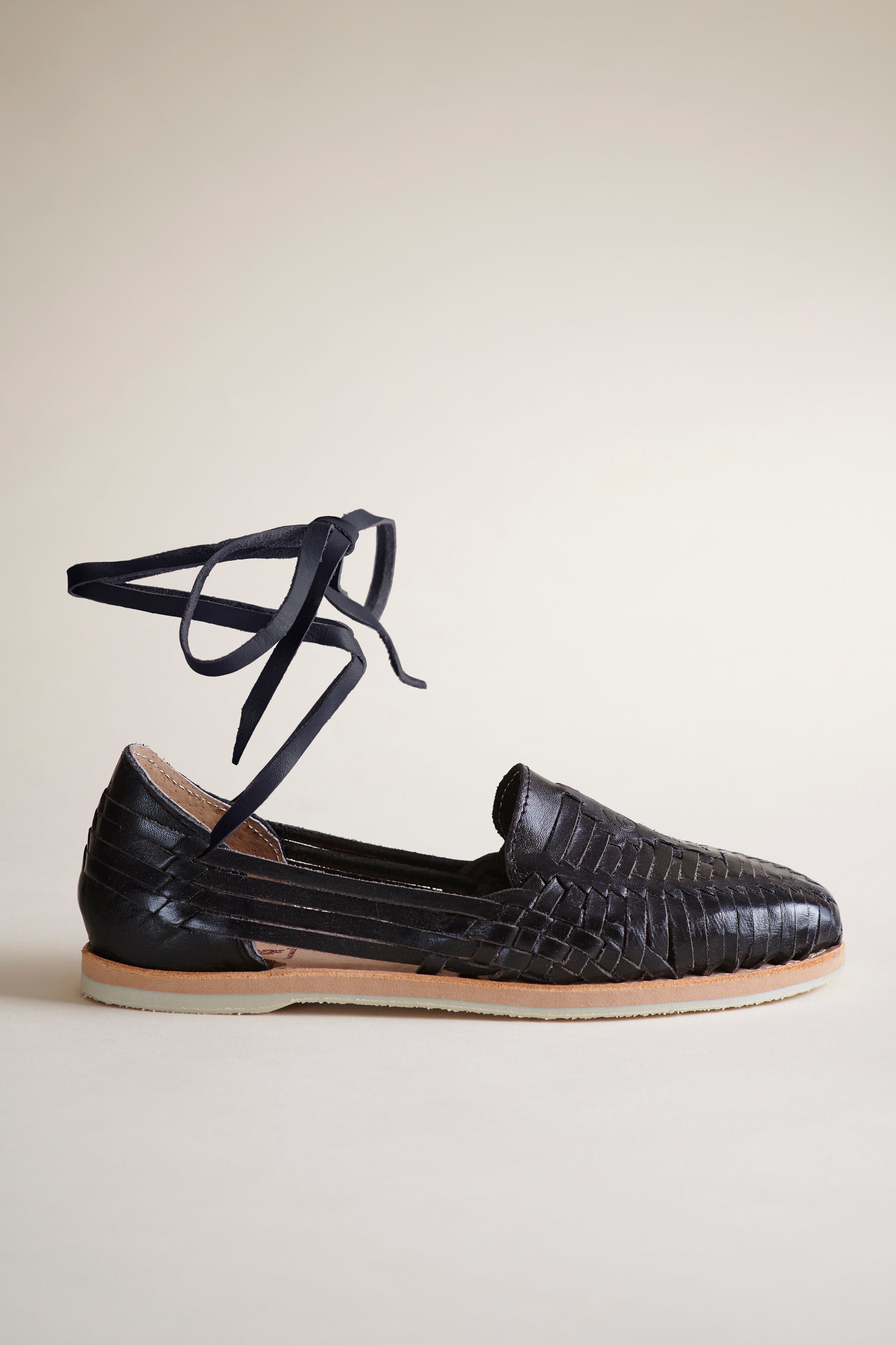 A single black braided leather flat with thin black lace-up ankle ties, shown in profile against a warm beige background.