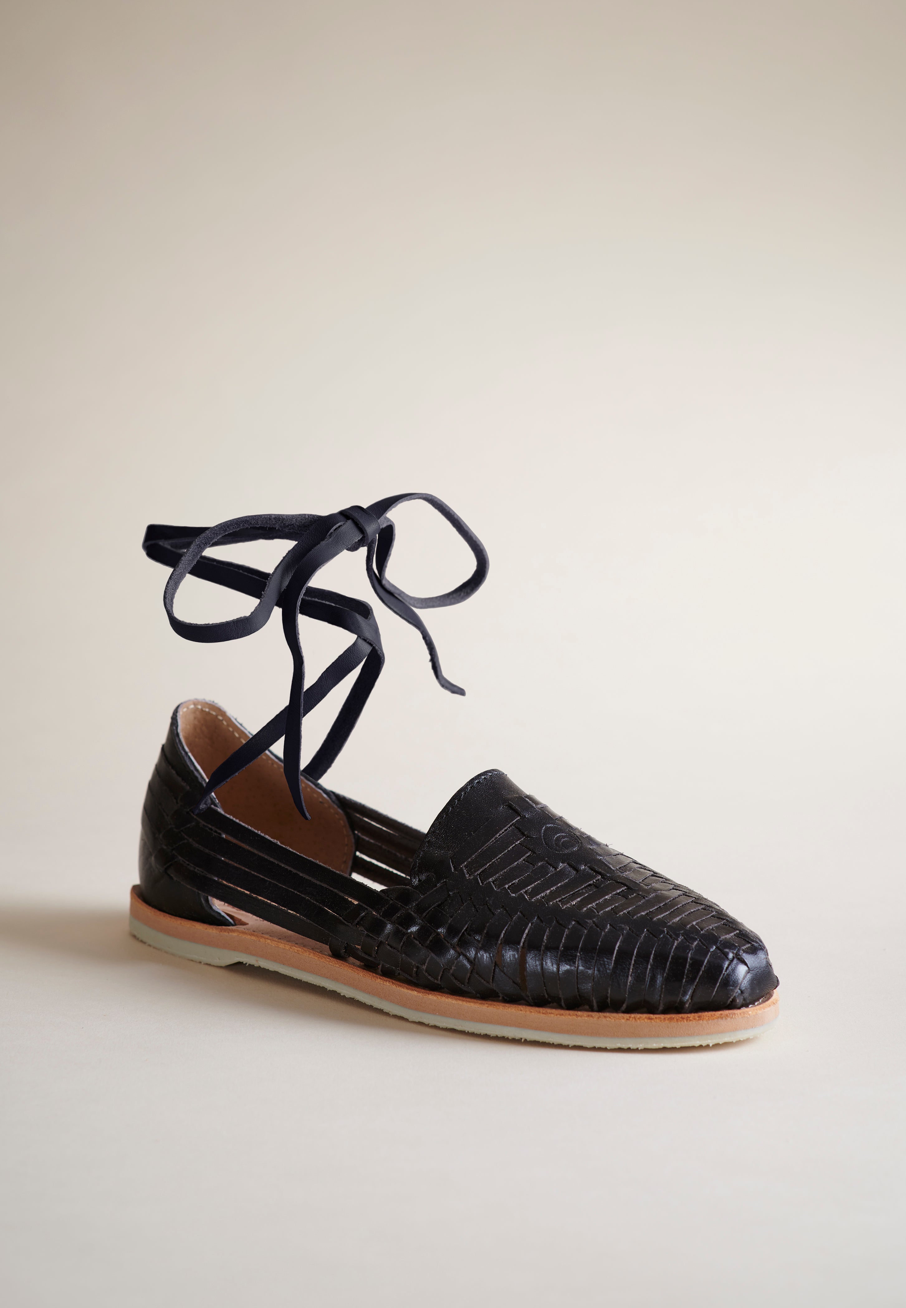 A single black leather flat with matching lace-up ankle ties, shown on a neutral beige background.