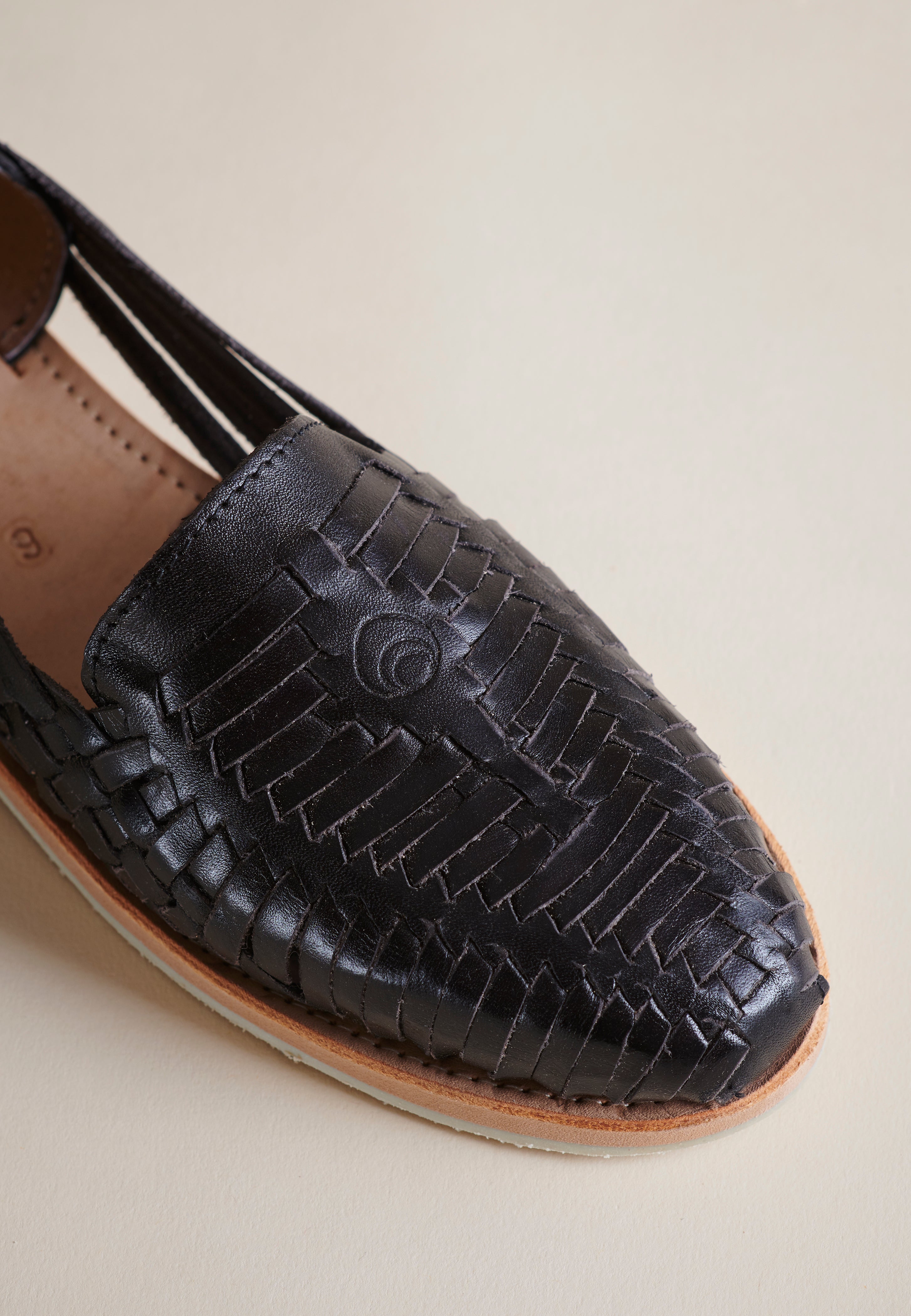 A close-up of a black woven leather loafer-style shoe with a tan sole, photographed on a plain warm beige background.