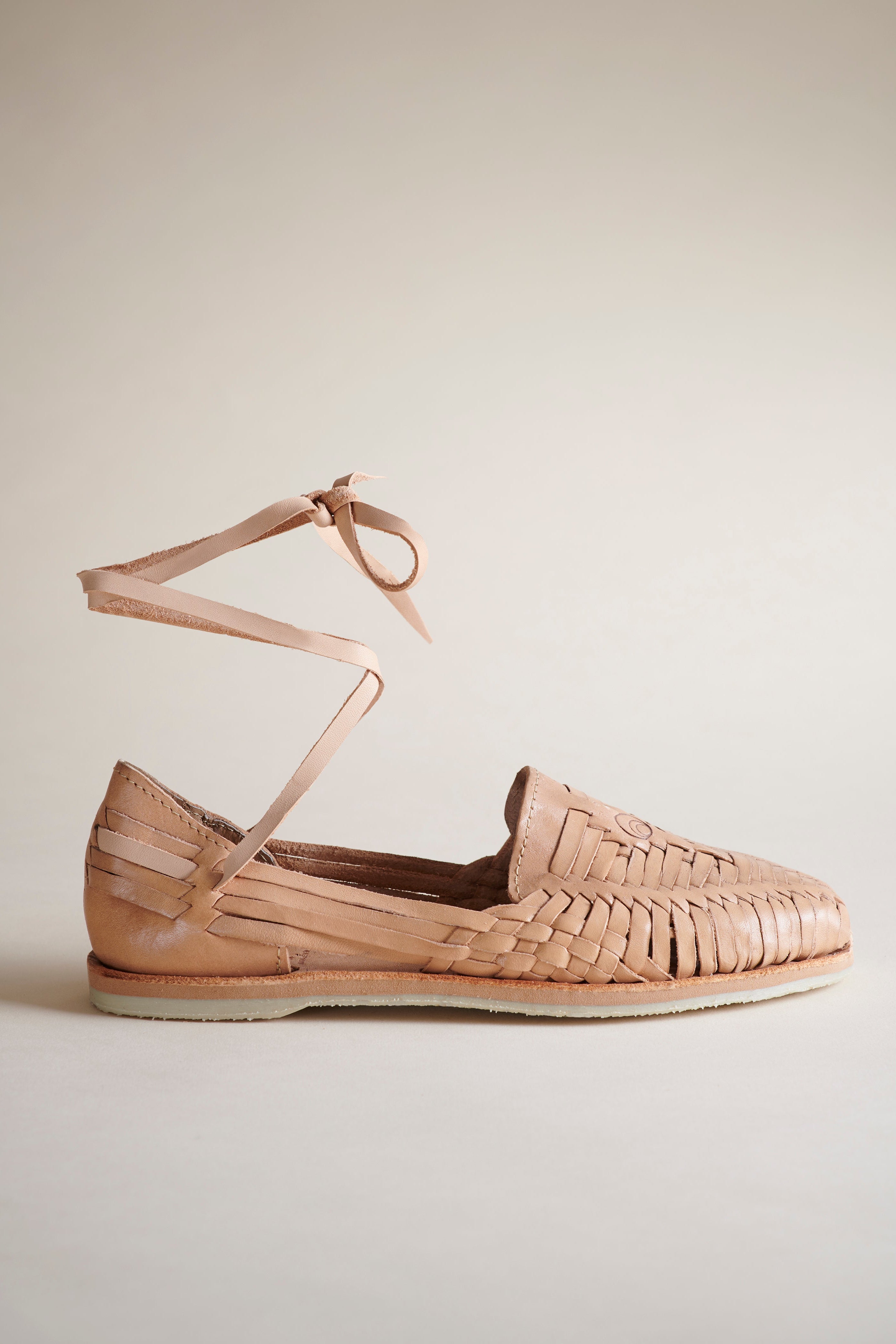 A single tan leather woven flat with an ankle tie, shown from a side profile against a neutral beige background.