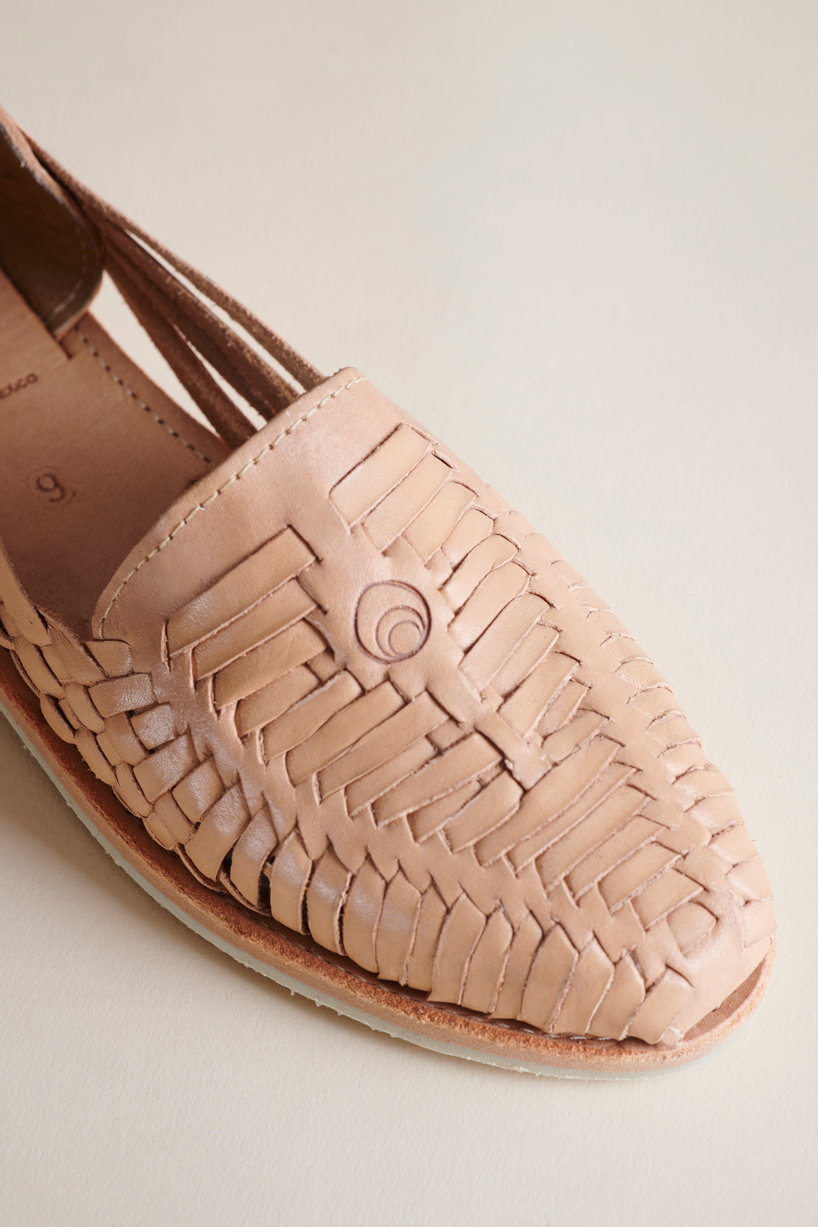 A close-up of a woven leather loafer in a light beige or nude color, showing the detailed basketweave texture and a spiral decorative detail on the toe, set against a plain light beige background.
