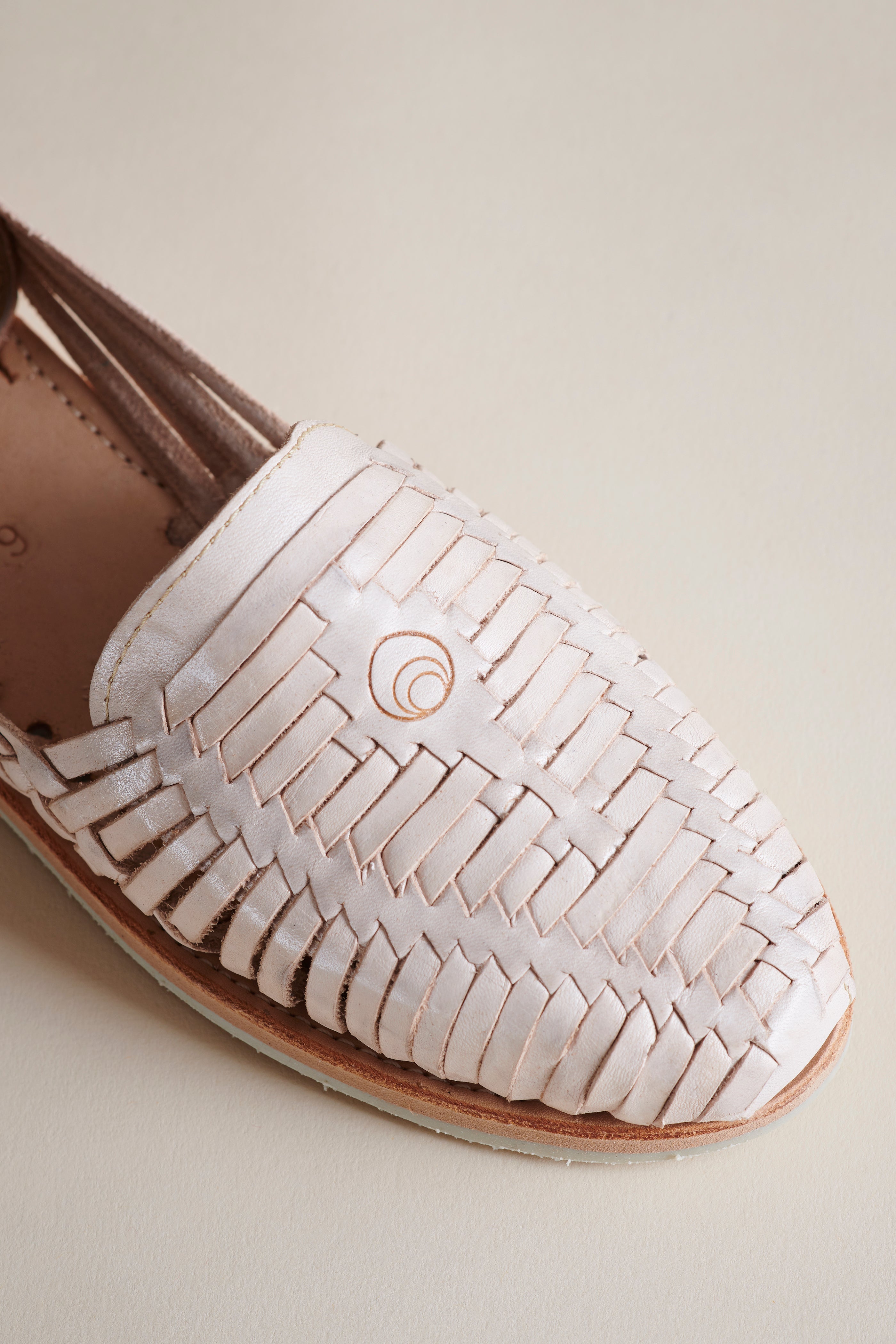 A close-up of a woven leather loafer in a light cream or off-white color, featuring a textured braided pattern and a small circular logo on the side, set against a solid light beige background.