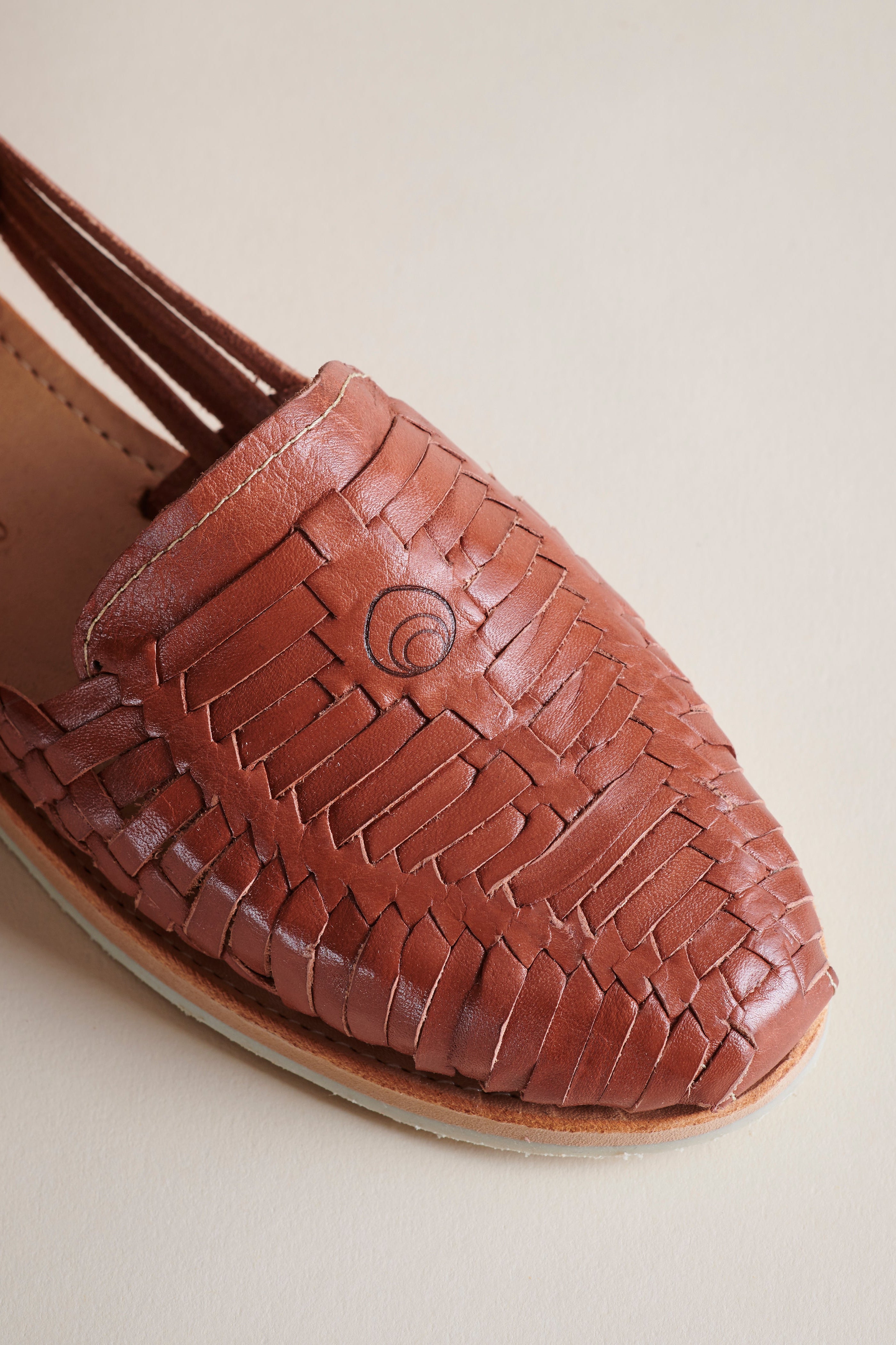 A close-up of a brown woven leather loafer-style shoe with a pointed toe and braided strap detailing, photographed on a plain light beige background.