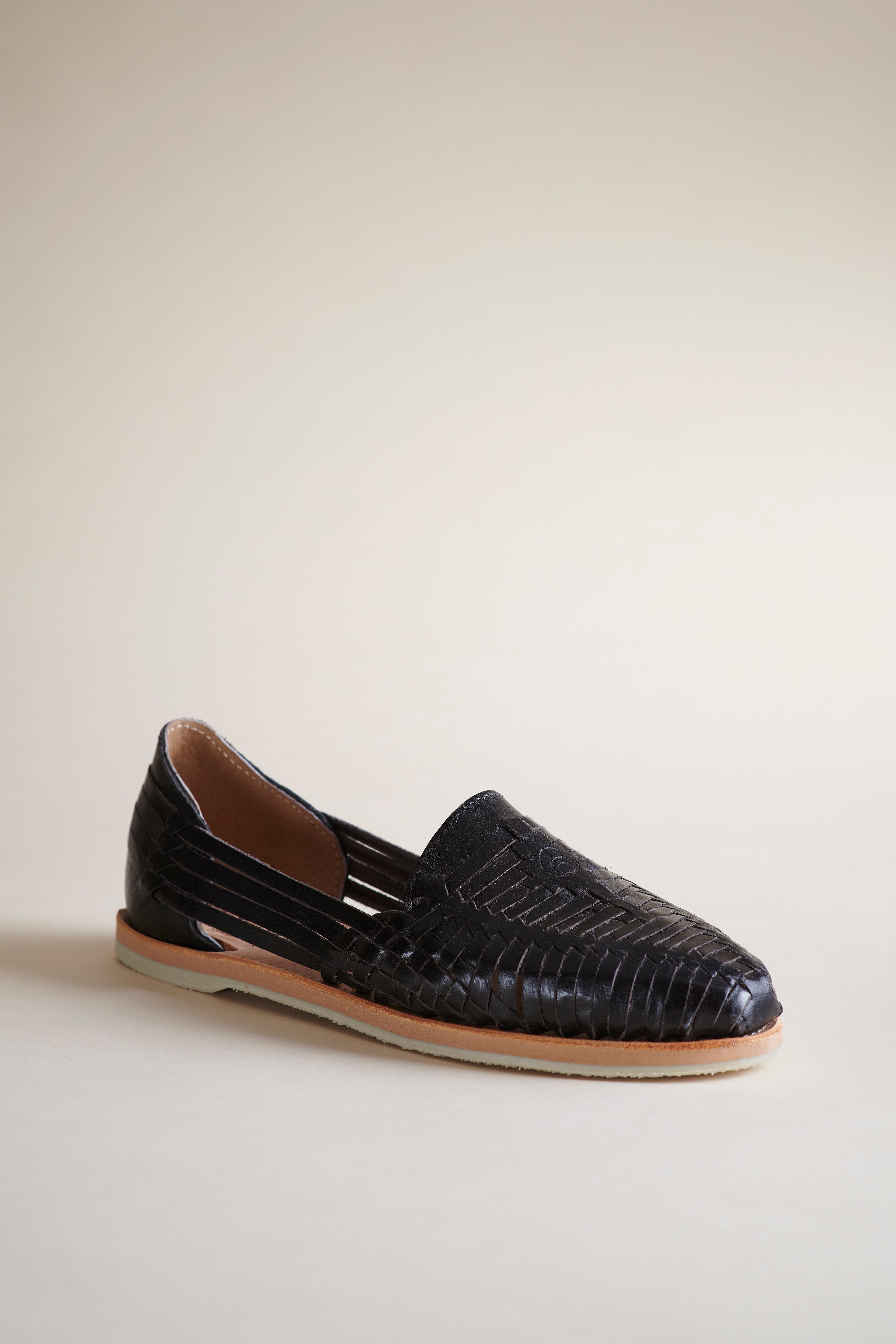 A single black braided leather slip-on flat with a tan sole, photographed from the side against a solid beige background.