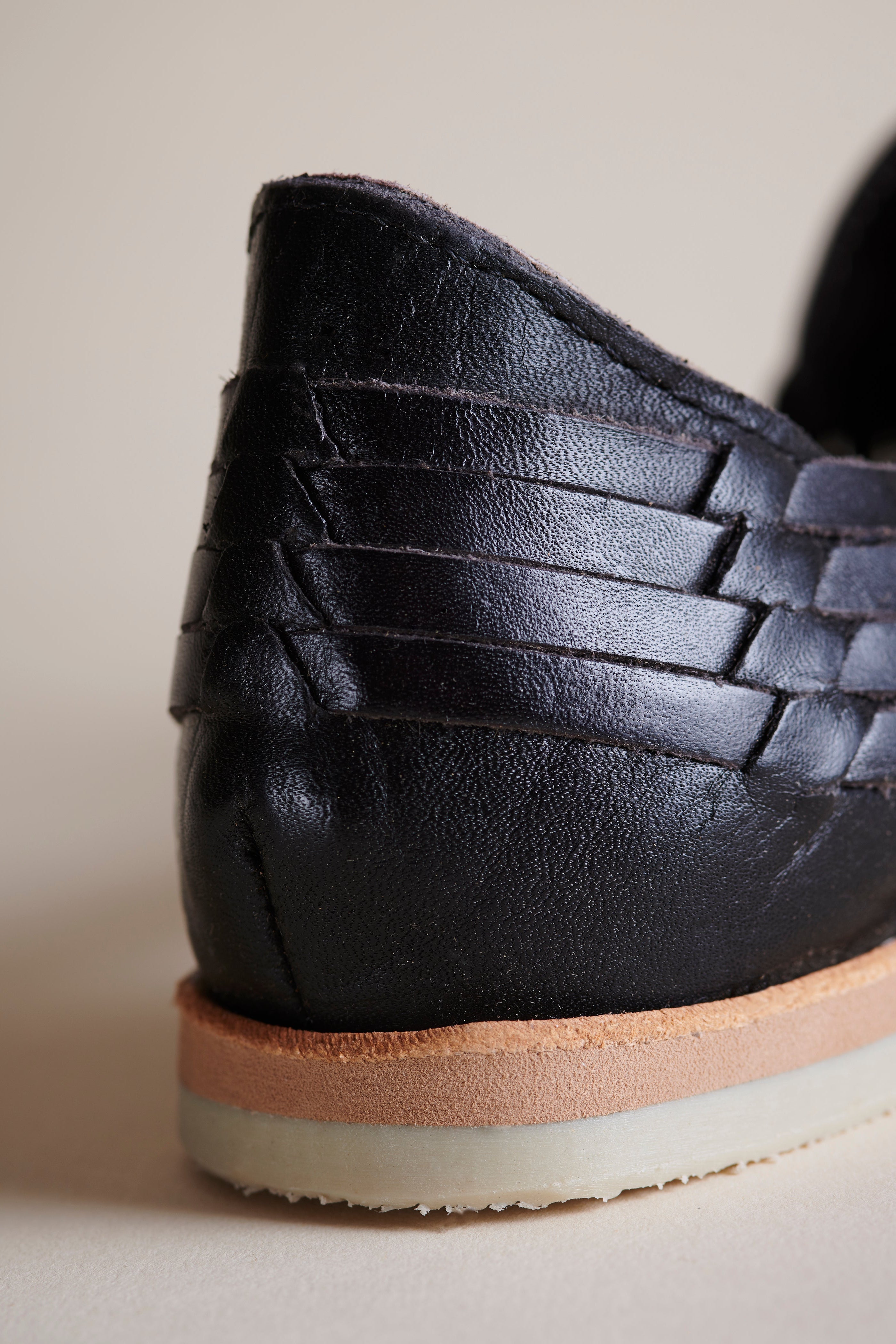 Close-up of the heel of a black leather shoe with a braided strap detail and a layered sole in tan and off-white, shown against a neutral beige background.