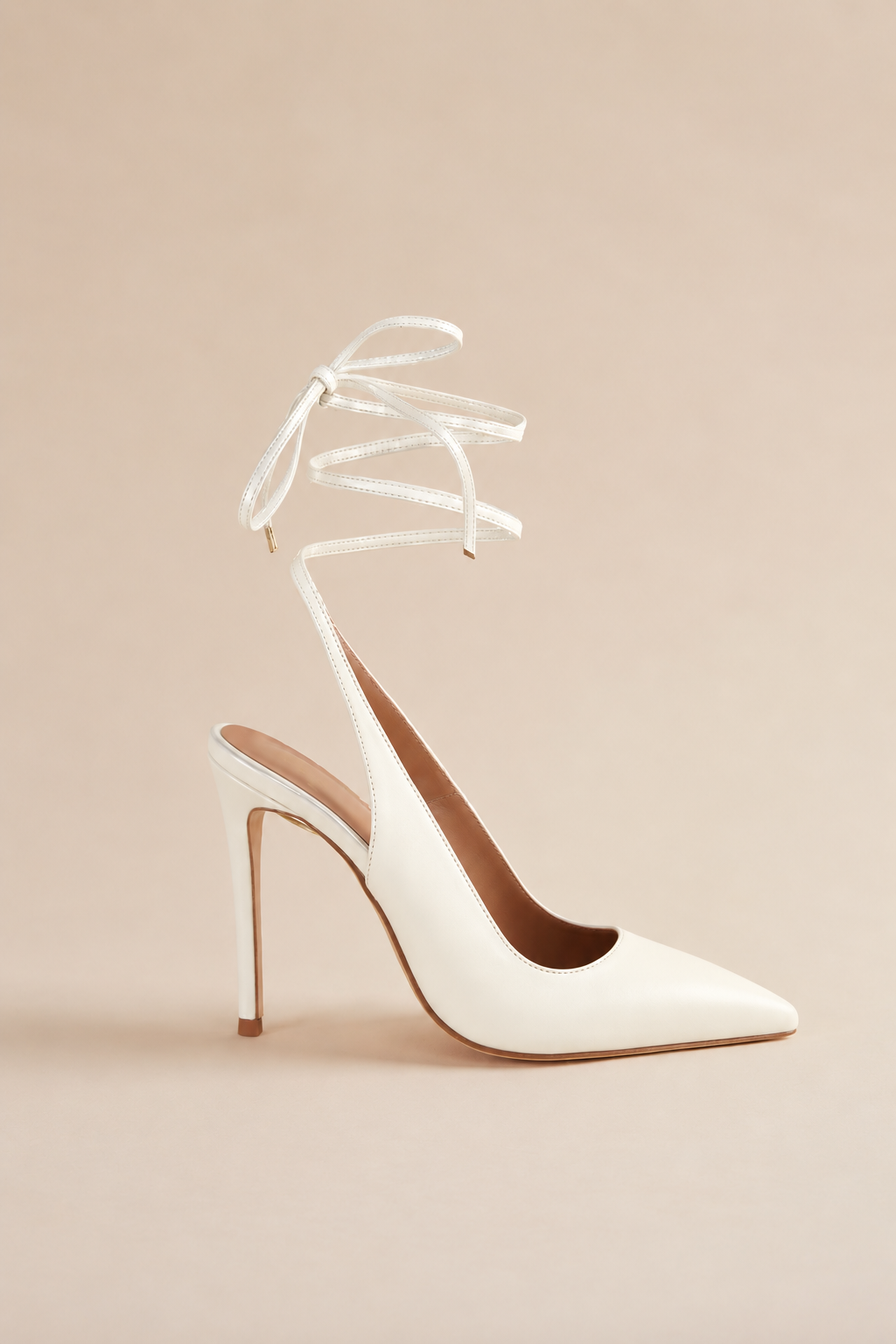 A single white pointed-toe slingback high heel with delicate wraparound ankle straps tied in a bow, shown against a solid beige background.
