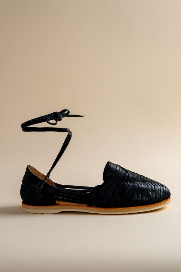 A single black leather Huarache flat shoe with a lace-up ankle tie against a beige background.