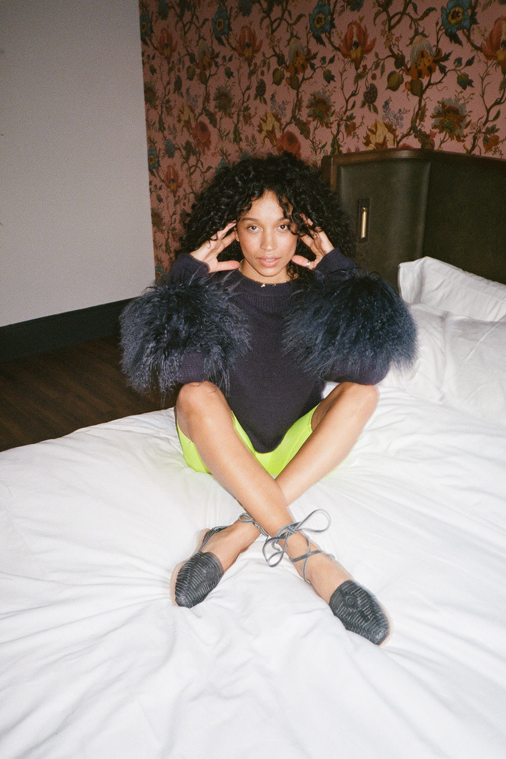 editorial image of a woman sitting on a hotel bed wearing a black shirt with feathers, green leggings, and black leather flats