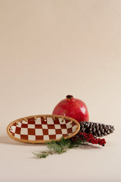 Brother Vellies Checkered Ceramic Dish in red & white checkers with a wicker border leaning up against a red fruit, acorn, and a pine leaf against a beige backdrop.